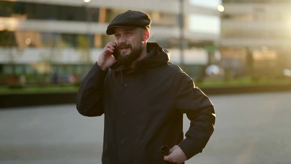 a Mustachioed and Bearded Man in a Cap and a Dark Jacket Makes a Phone Call Against the Background