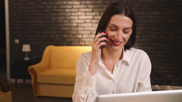 Smiling Pretty Caucasian Woman Talking on Phone Using Laptop at Home or Office