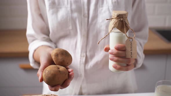 Potato Milk In Glass Bottle In One Hand, Two Potato Tubers In The Other. Vegan Plant Based Milk