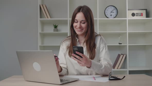 Smiling Young Woman Customer Holding Credit Card and Smartphone Sitting at Home Office