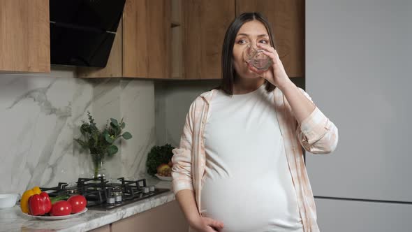 Pregnant Woman Caresses Belly and Drinks Water in Kitchen