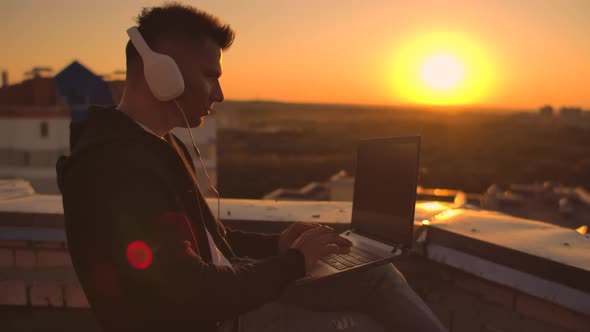 A Man in a Hoodie Sits on the Roof and Listens To Music with Headphones Typing on a Laptop Keyboard.