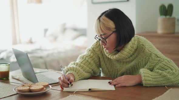 Pensive Asian woman in eyeglasses writing something in notepad from laptop
