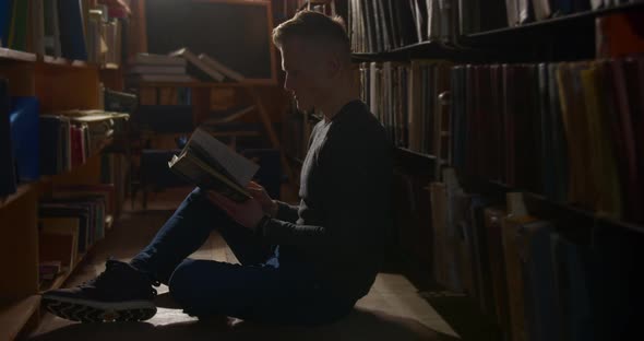 A Young Guy Is Sitting on the Floor Between the Bookshelves and Leafing Through a Book. 