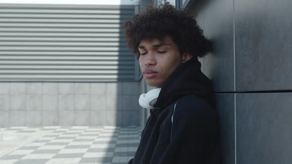 A Troubled Teenager with a Nose Piercing in a Black Hoodie and Headphones Sits Leaning Against a