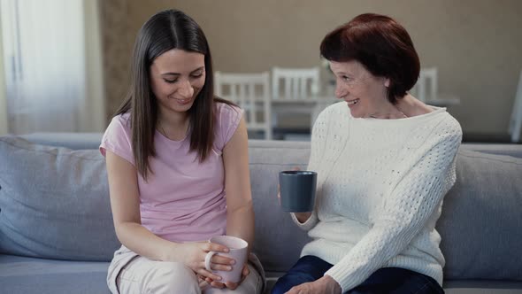 Mother and Daughter are Sitting on the Sofa at Home and Drinking Tea