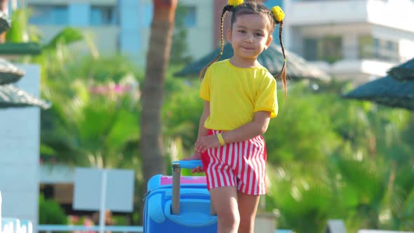 Little Cute Smiling Girl Is Walking with a Suitcase on a Resort Beach.