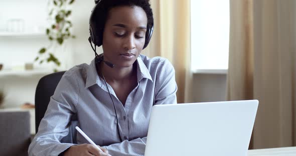 Focused Young African Student Wearing Wireless Headphones, Studying on Online Courses, Using Paper