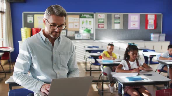 Portrait of happy caucasian male teacher in classroom with children using laptop looking at camera