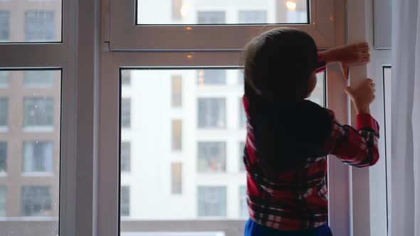 Boy Child in a Red Plaid Shirt Stands at the Window Where Snows in Winter