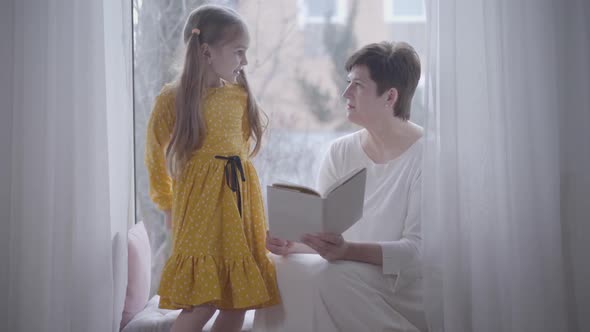 Pretty Caucasian Girl Coming To Grandmother Sitting on Windowsill with Book. Smiling Granddaughter