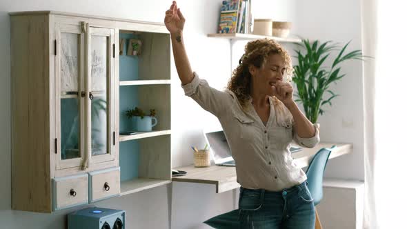 Curly blonde young adult woman dancing at home in living room with office workplace background