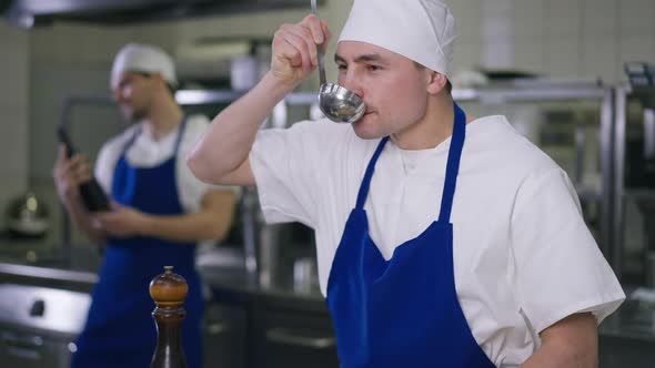 Caucasian Chef Tasting Soup Adding Salt to Dish Standing in Kitchen with Cook Preparing Food at