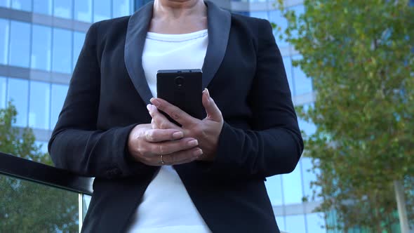 A Middle-aged Businesswoman Looks at a Smartphone in an Urban Area - a Windowed Office Building