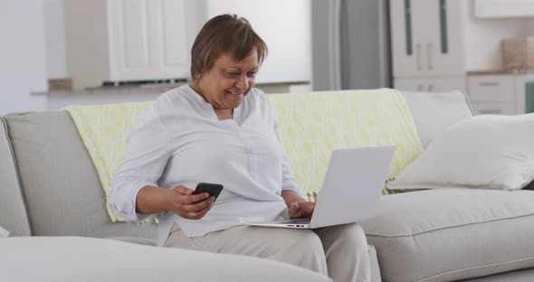 Happy african american senior woman sitting on couch using smartphone and laptop, smiling