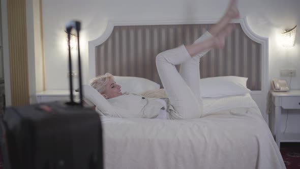 Joyful Blond Caucasian Woman Falling Back on Soft Luxurious Bed in Hotel Room and Smiling