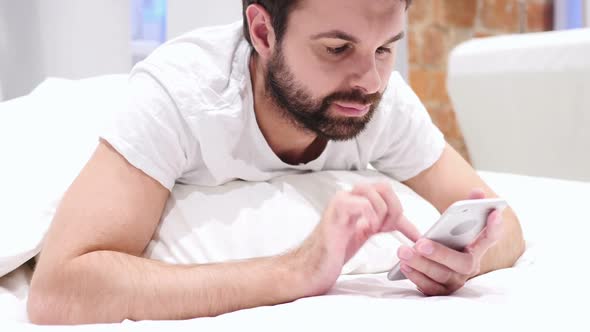 Lying Man on Stomach in Bed Browsing on Smartphone