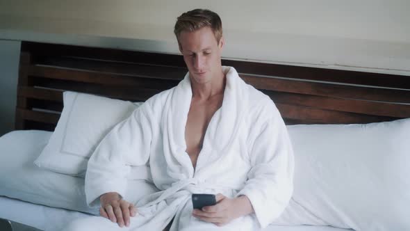 Handsome Blond Guy in White Bathrobe Use Mobile Phone and Answers Calling in Bed