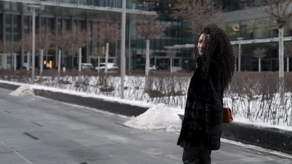 A Curlyhaired Woman in a Fur Coat Walks Through a Modern City Enjoying a Beautiful Day