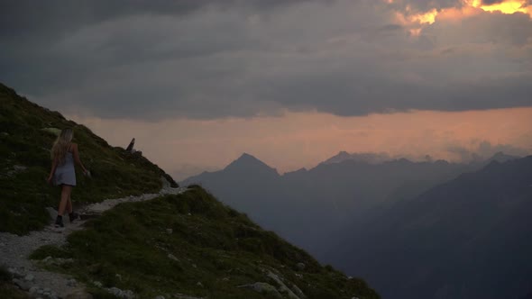 Very cinematic footage of a blonde girl in a white dress walking on an exposed mountain path towards