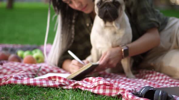 Portrait of a Girl Laying on Plaid on Lawn in a Park and Making Notes While Little Pug Sitting Next