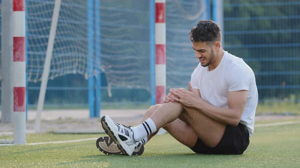 Unhappy Injured Middle Eastern Footballer Sitting on Grass of Soccer Field Against Goal Holding Knee