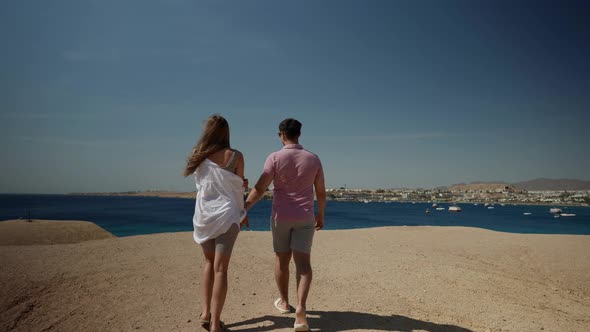 a Man in a Pink Tshirt and a Woman with Long Hair in a White Shirt Hold Hands and Walk to the Sea