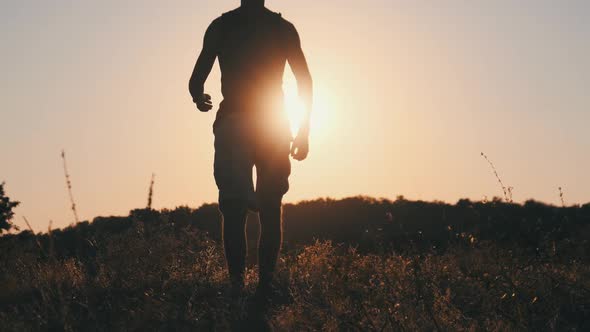 Silhouette of Young Man Walking in Field To Setting Sun and Raising Hands Up