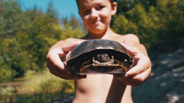 Boy Holds Turtle in Arms and Smiles on Background of River with Green Vegetation