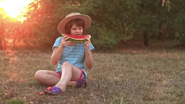 Funny Little Girl Smile and Eat Watermelon Slice Sitting on Grass with Crossed Legs in Summer Park