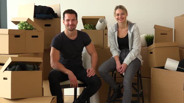 A Happy Moving Couple Sits on Chairs and Smiles at the Camera in an Empty Apartment