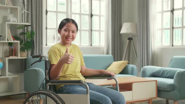 Asian Kid Girl Sitting In A Wheelchair Smiling And Showing Thumbs Up In Living Room