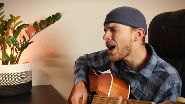 Male singer sings song and plays guitar at home in front of laptop, learns new music composition.