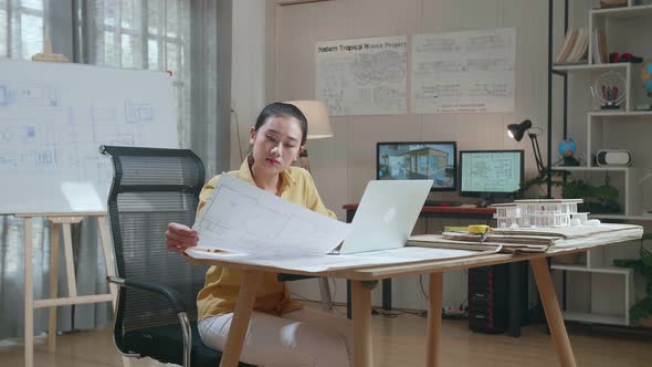 Asian Woman Engineer Typing On A Laptop And Looking At Blueprint Before Walking Out Of The Office