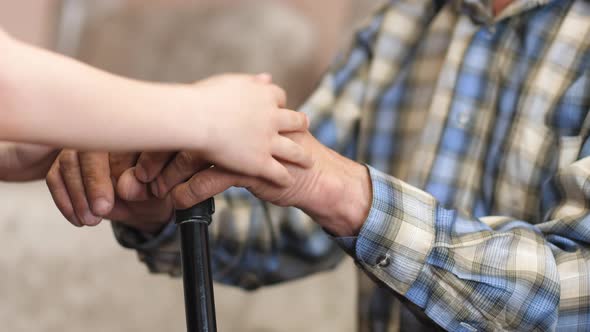 The hands of a child hug and stroke the arms of an elderly man sitting with a walking stick. Caring 