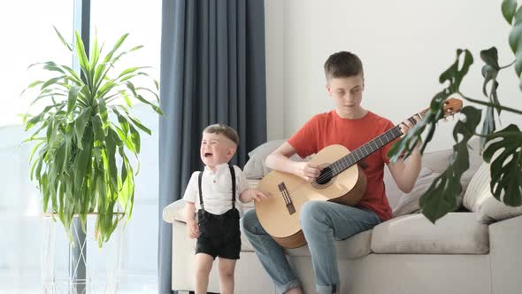 A Little Boy is Crying and His Brother is Playing the Guitar While Sitting on the Couch