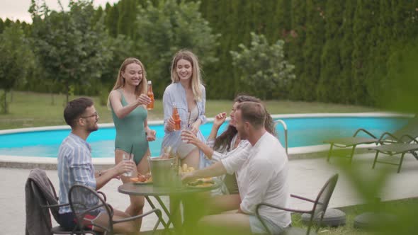 Group of happy young people cheering with drinks and eating fruits by the pool in the garden