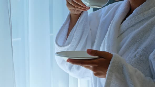 A beautiful young dark-skinned mixed-race woman stands by the window in the hotel and drinks coffee.