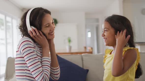 Happy hispanic mother wearing headphones listening to music and daughter sitting on sofa