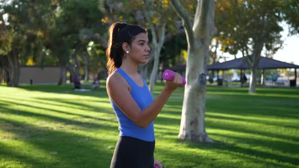 A young woman doing a dumbbell workout in the park performing bicep curls to build arm muscle and st