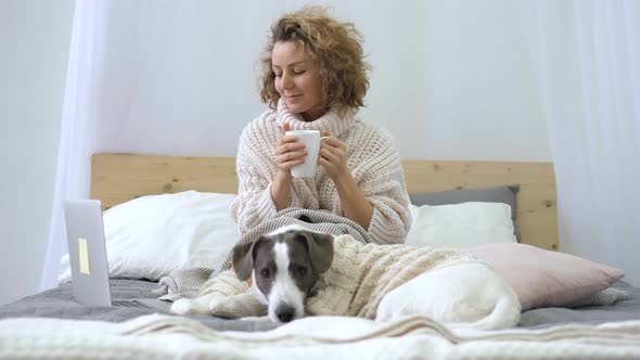 Happy Casual Woman Working On A Laptop Sitting On Bed With Dog