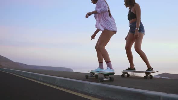 Two Girlfriend Girls in Shorts and Sneakers Ride Skateboards on the Slope Against the Beautiful Sky