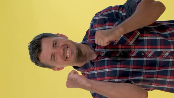 Excited Man in Checkered Shirt Raising Fists in Cheer on Yellow Background