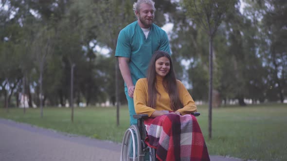 Wide Shot of Caucasian Man Strolling with Disabled Woman on Wheelchair in Summer Park and Stopping