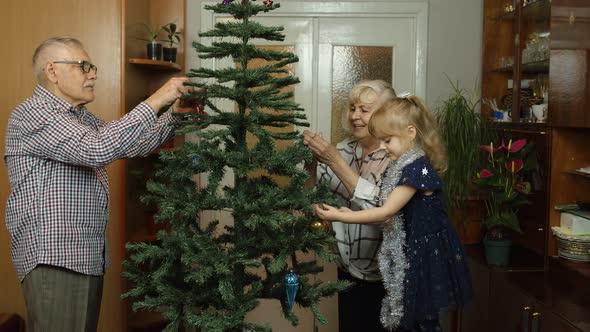 Kid Girl with Senior Grandmother and Grandfather Decorating Artificial Christmas Tree with Toys