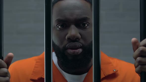 Aggressive Afro-American Criminal Standing in Jail With Hands on Prison Bars