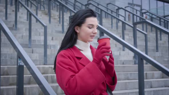 Profile Portrait of Charming Caucasian Lady in Red Coat and Gloves Drinking Coffee in City. Young