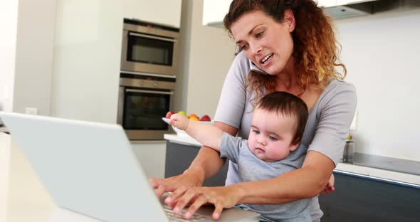 Mother sitting with baby boy on lap using laptop and talking on phone