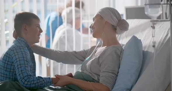 Teen Son Talking and Holding Hand of Mother in Headscarf Laying in Hospital Bed After Chemotherapy