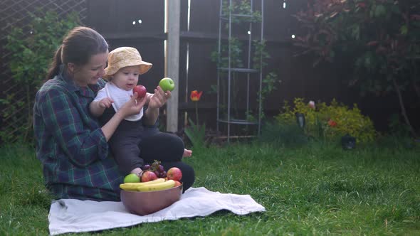 Happy Young Cheerful Mother Holding Baby Eating Fruits On Green Grass